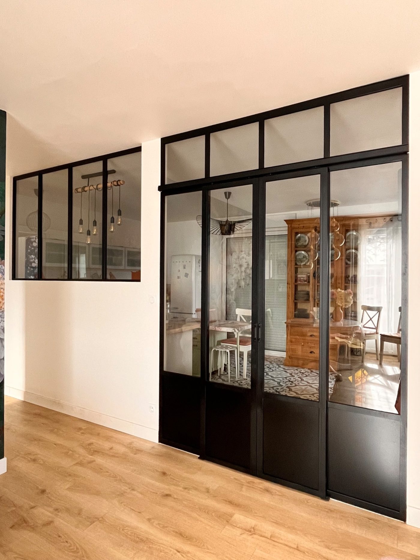 Verrière d'atelier noire en acier avec portes coulissantes vitrées et imposte, séparant le salon de la cuisine, avec une seconde verrière fixe sur le mur.