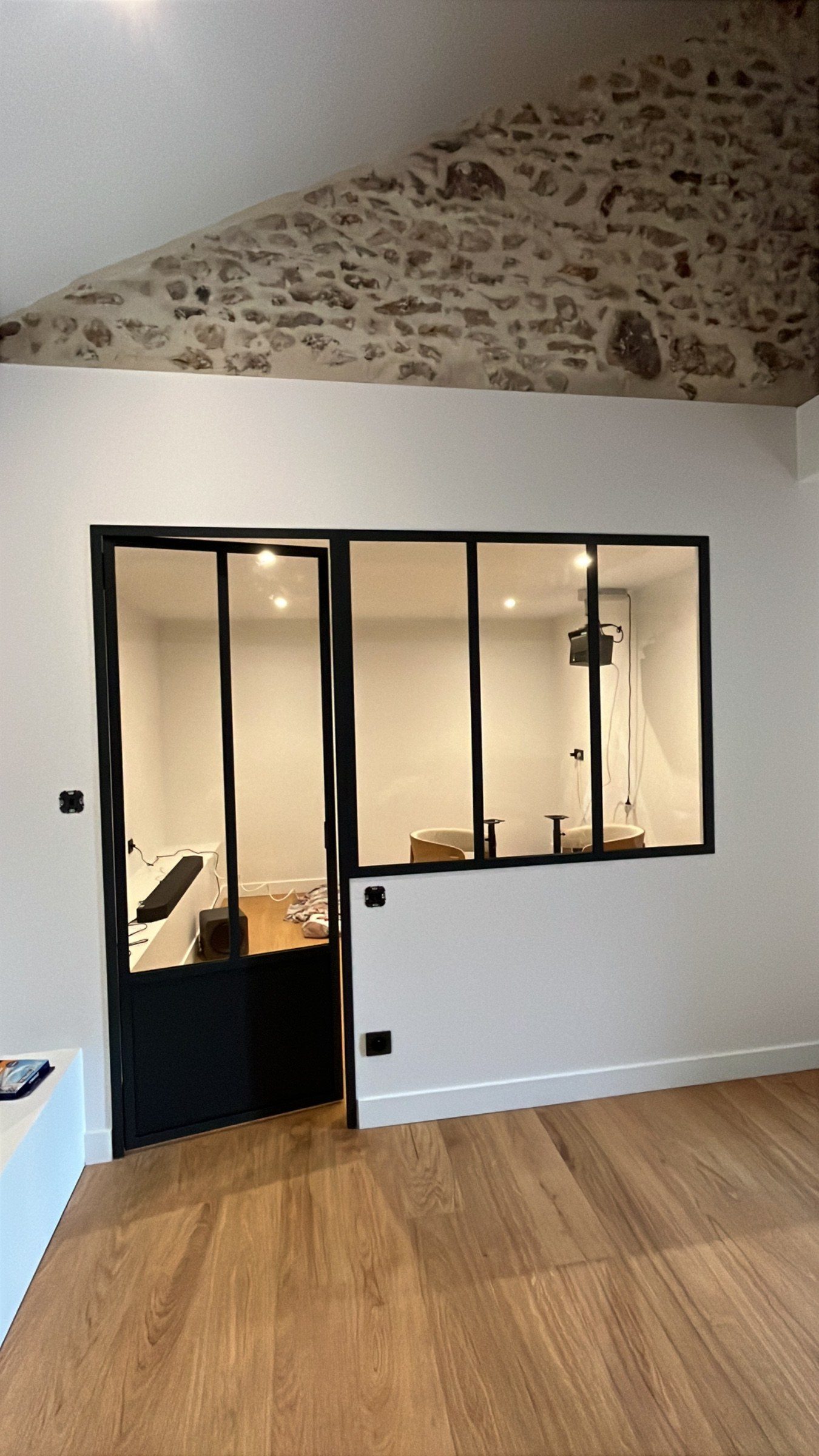 Verrière en acier noir avec porte pleine en partie basse, posée dans un bureau sous un plafond en pierre apparente, parquet au sol.